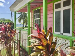 Pink Colored Typical Barbados Chattelhouse With White And Green Decor Color Along Porch Rails And Leafes Of Plants With View To The Sea
